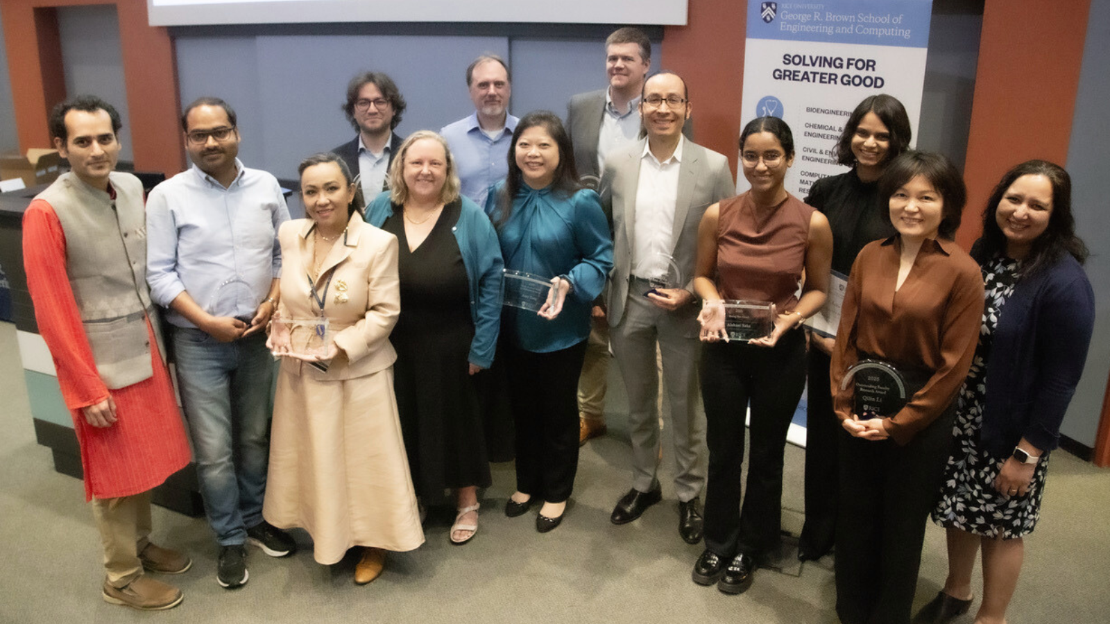 Group of professionals, some holding awards pose together in front of a banner for the George R Brown School of Engineering and Computing.