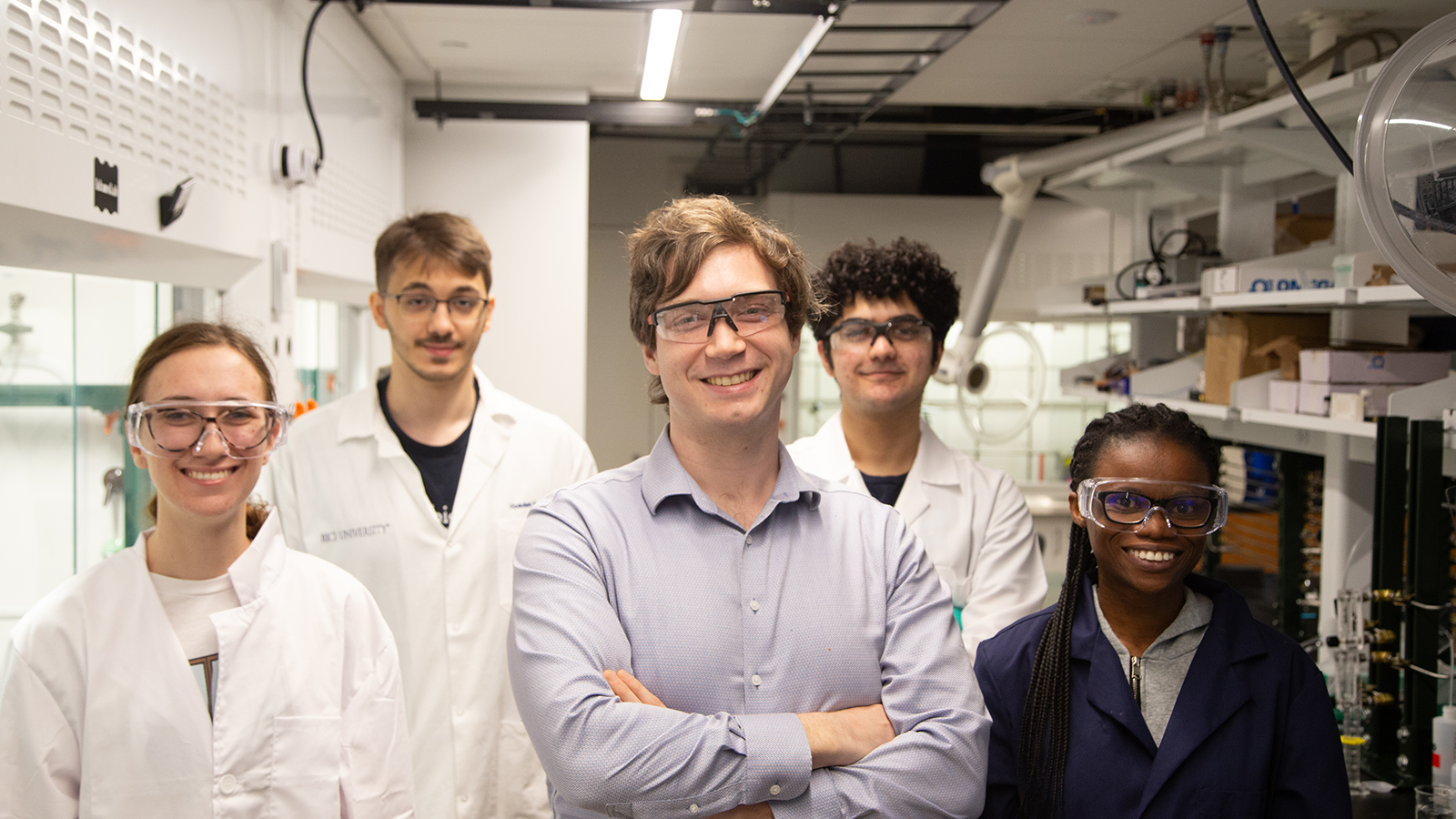 Jason Adams poses with four members of his group in a lab setting. Adams wears a button down shirt; other team members wear lab coats. All of the individuals in the photo are wearing glasses and/or safety goggles.