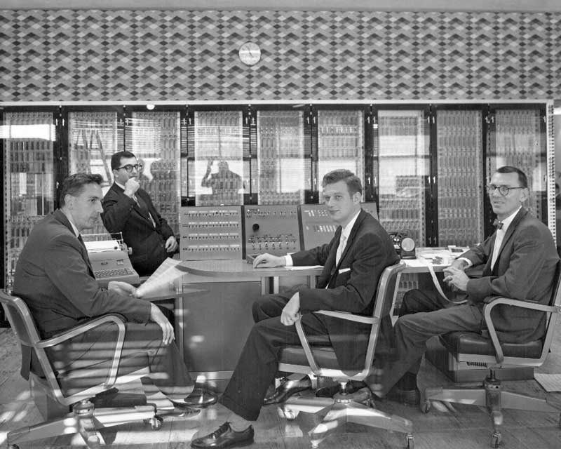 Four men (from left to right): John Kenneth Iliffe, Martin Graham, Zevi Salsburg, and John Kilpatrick sitting in front of the R1 (Rice University Computer Project), located in Abercrombie Laboratory at Rice University.